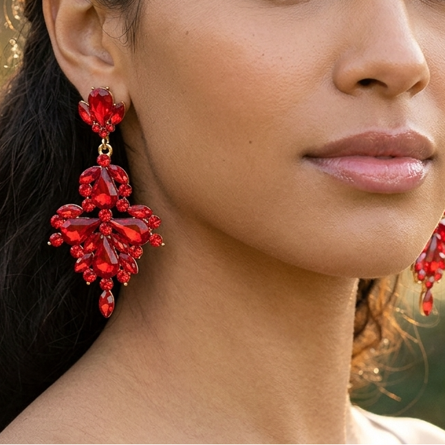 A close-up portrait of a woman wearing dramatic, chunky red crystal drop earrings against a softly blurred outdoor background.