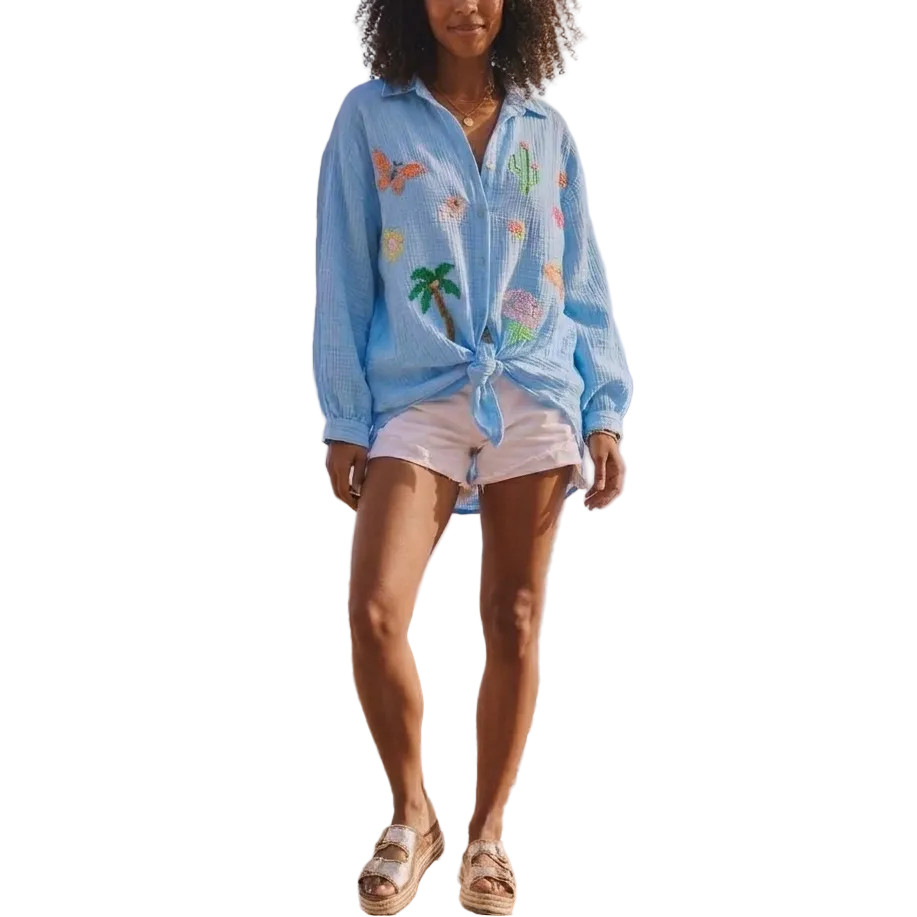 Woman standing on a rocky beach wearing a light blue embroidered shirt and white shorts.