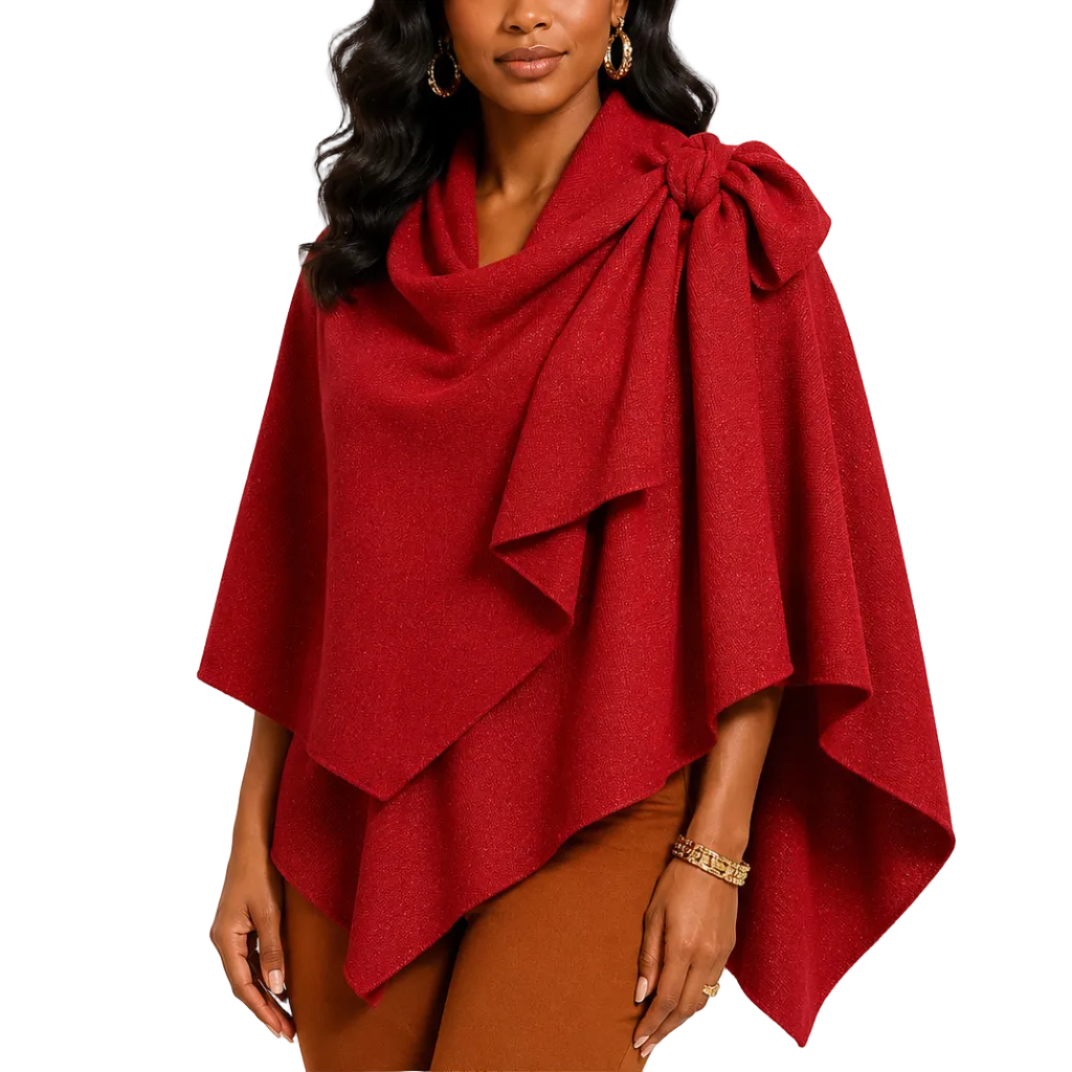 A woman modeling a deep red draped wrap poncho with a large bow detail at the neckline, styled with brown pants against a plain white studio background.