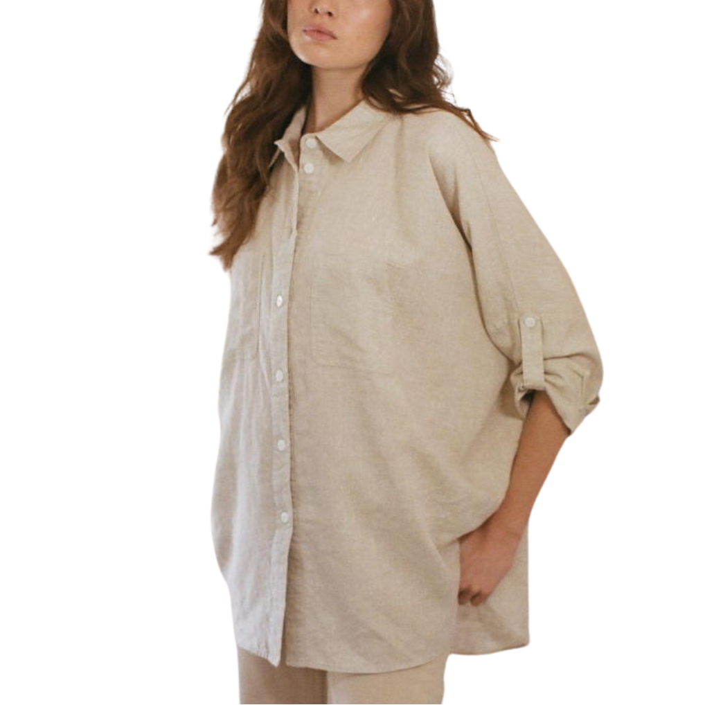 A woman wearing a beige button down collar shirt made of linen, with long sleeves and front pockets, standing against a white background.
