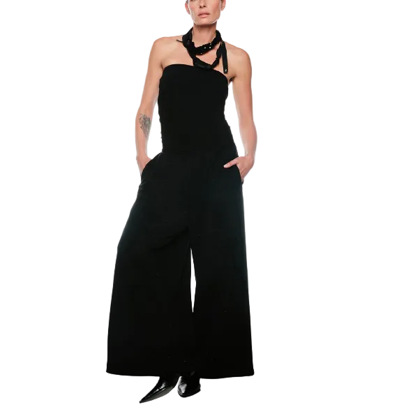 A model stands against a plain white studio background wearing a black halter-style wide-leg jumpsuit with a chunky black collar necklace and black pointed shoes.