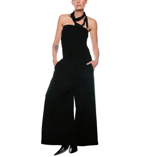 A model stands against a plain white studio background wearing a black halter-style wide-leg jumpsuit with a chunky black collar necklace and black pointed shoes.