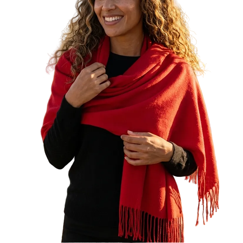 A smiling woman with curly brown hair stands outdoors wearing a bright red shawl or wrap over a black top, with a vineyard and sea in the blurred background.