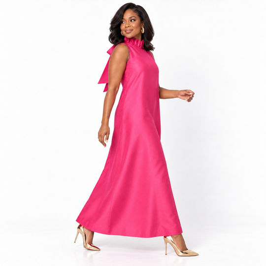 A woman modeling a bright pink sleeveless maxi dress with a high ruffled neckline and a large bow detail on the left shoulder, paired with metallic gold high-heeled sandals against a plain white studio background.