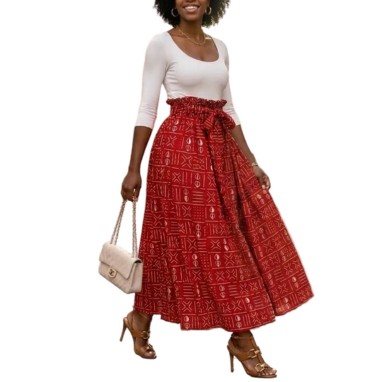 A woman stands on a cobblestone street wearing a white fitted top and a full red midi skirt with a white geometric pattern and a tied waist. She carries a small white handbag and wears brown heeled sandals.