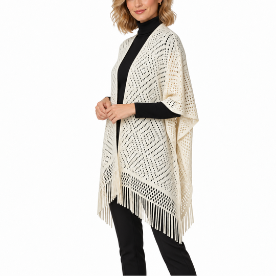 A woman modeling a cream open-knit fringed poncho over a black turtleneck and black pants, photographed against a plain white studio background.