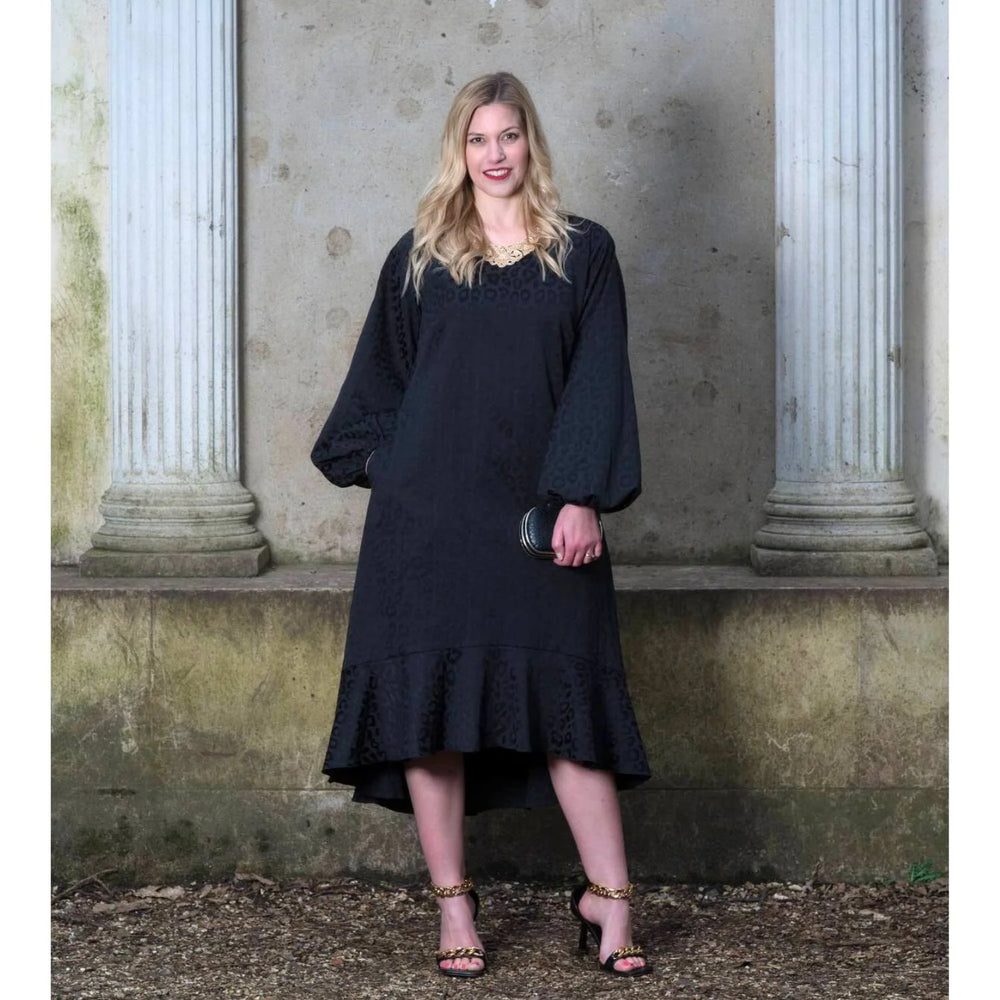 A woman standing in front of a classical columned background, wearing a long black dress with three-quarter sleeves and a V-neck.