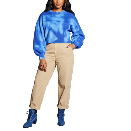 Woman in blue sweater and beige pants standing on a rooftop with cityscape background