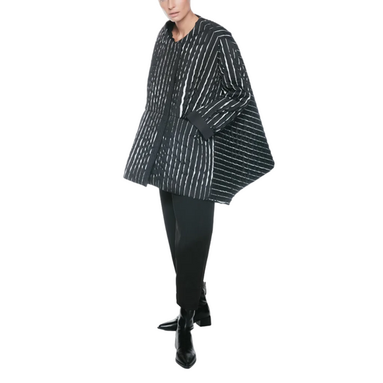 A model wearing a black and white vertically striped cape-style jacket, black trousers, and black ankle boots, standing against a plain white studio background.