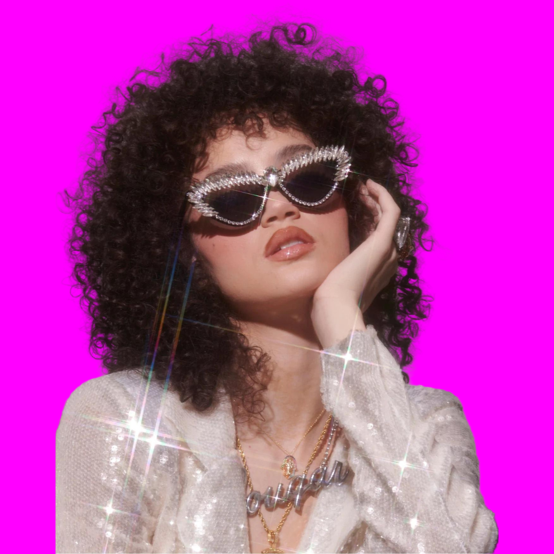 Person with curly hair wearing sparkling sunglasses and a sequin outfit against a plain background