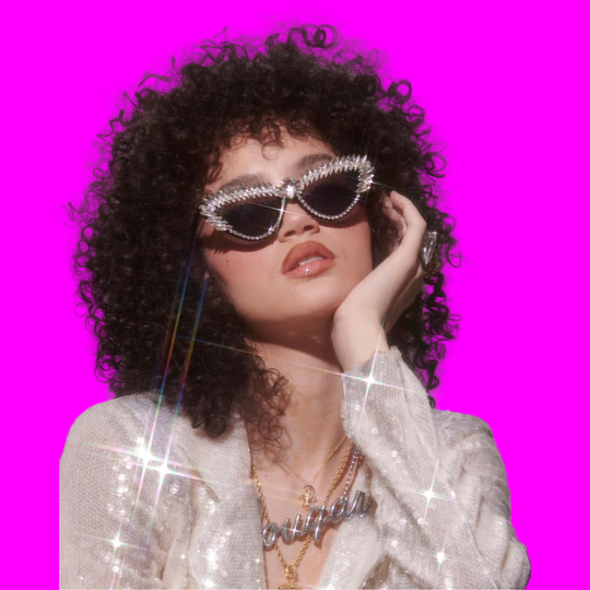 Person with curly hair wearing sparkling sunglasses and a sequin outfit against a plain background