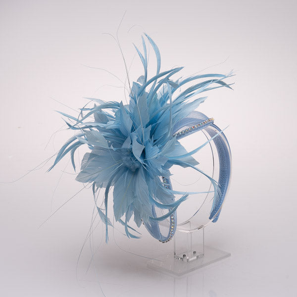 A light blue feather fascinator headpiece with a matching feathered headband and clear rhinestone trim, displayed on a clear acrylic stand against a white background.