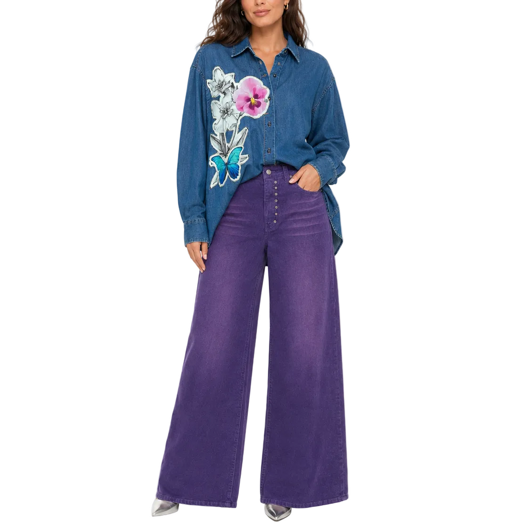 A woman modeling a blue long-sleeve button-up shirt with a colorful floral and butterfly graphic on the front, paired with wide-leg purple jeans, against a plain white background.
