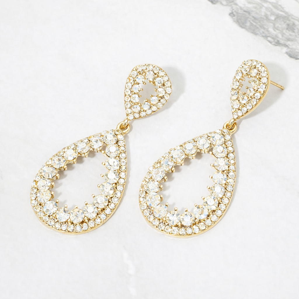 A pair of gold teardrop-shaped drop earrings adorned with clear rhinestones, displayed on a light gray marble surface.