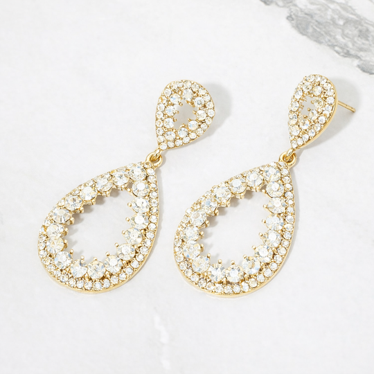 A pair of gold teardrop-shaped drop earrings adorned with clear rhinestones, displayed on a light gray marble surface.