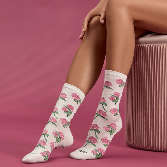 A person seated on a pink ribbed stool wearing white crew socks with an allover pink flower and green leaf pattern, against a solid pink studio backdrop.