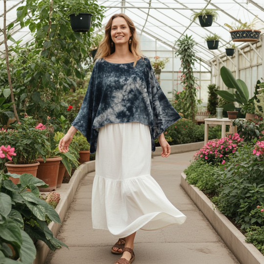 Woman in a tie-dye top and white skirt walking through a greenhouse filled with plants and flowers.