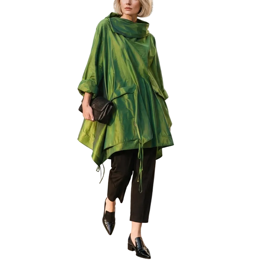 A woman stands outdoors in a modern urban setting wearing a shiny olive green poncho-style top, black cropped pants, black pointed shoes, and holding a black clutch bag.