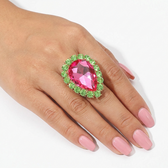 Ring with pink gemstone and green accents on a hand with pink nail polish against a white background