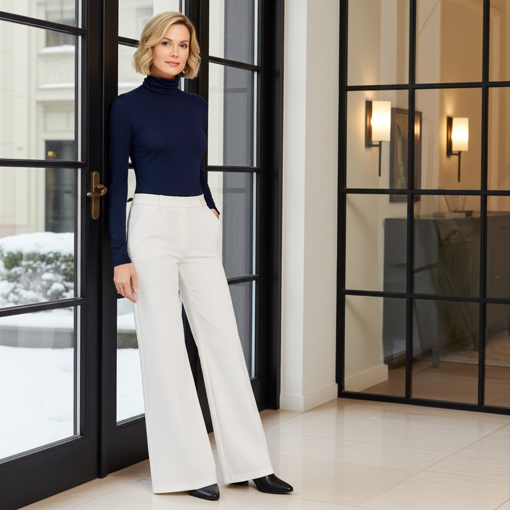 Woman in navy turtleneck and white pants standing in a modern interior setting.