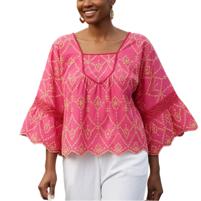 Woman in a pink blouse and white pants standing in a greenhouse filled with plants and flowers.