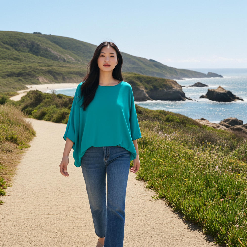 Woman in teal top and jeans walking on a path by the ocean with green hills in the background