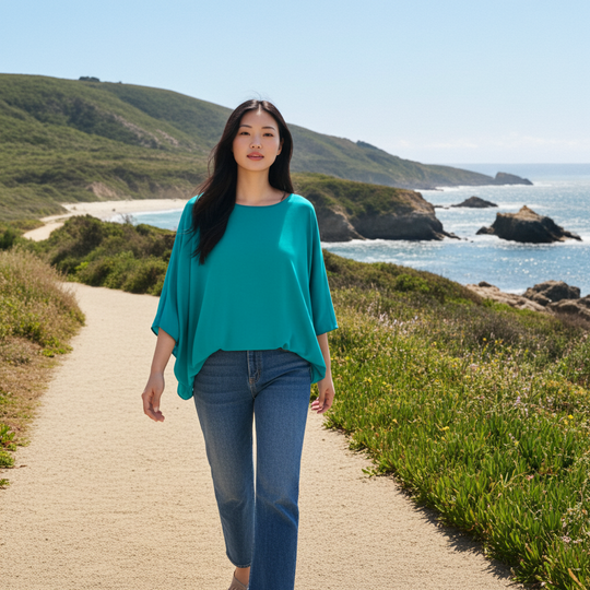 Woman in teal top and jeans walking on a path by the ocean with green hills in the background