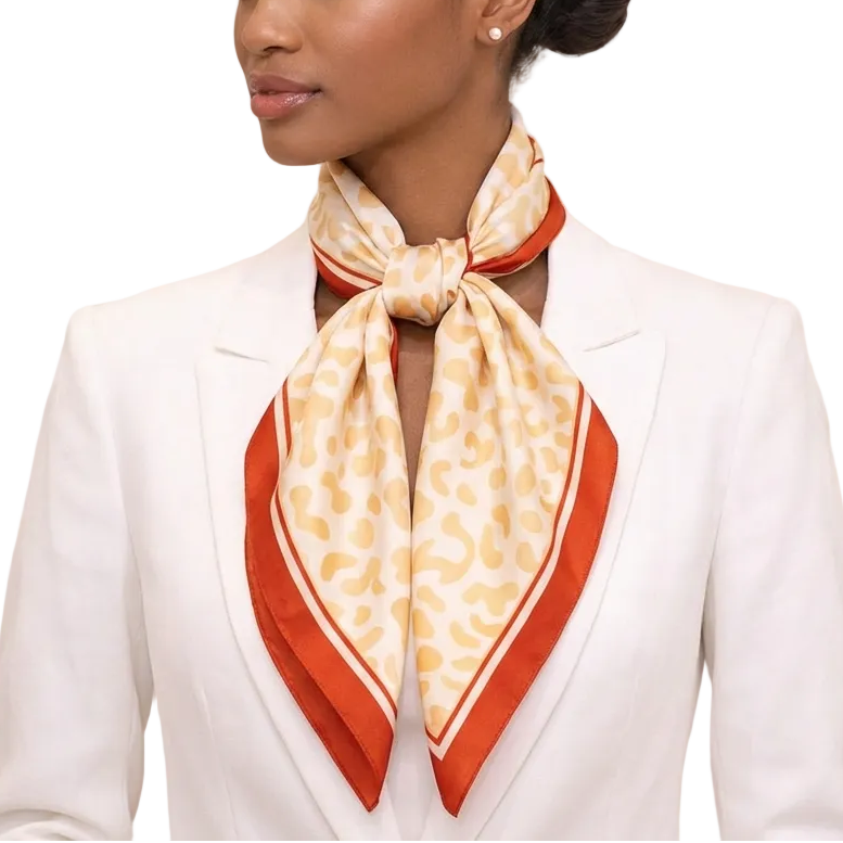African American woman in a white blazer wearing a beige leopard-print silk scarf with red border, tied in a front bow, shown in a soft high-end boutique setting.