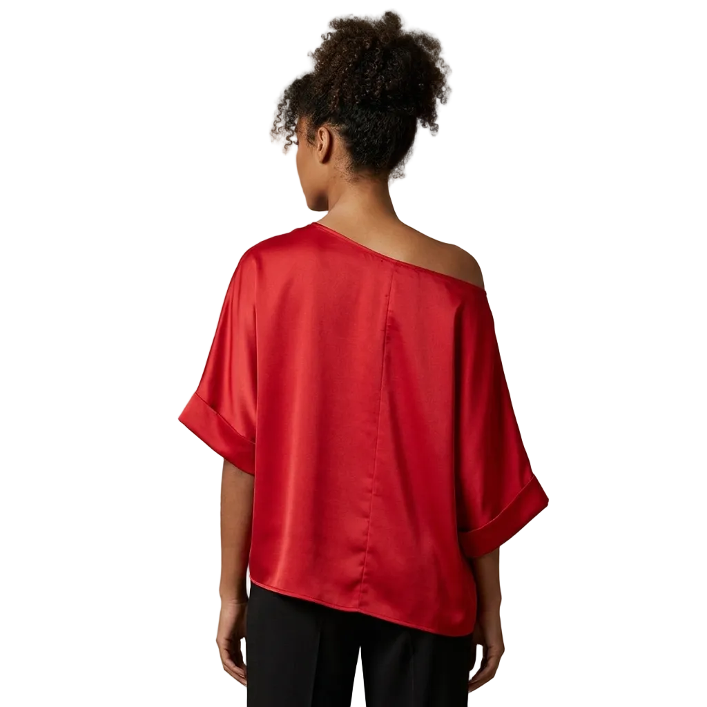 A woman viewed from the back wearing a bright red satin-like off-the-shoulder top with wide elbow-length sleeves and black pants, standing against a plain warm beige studio background.