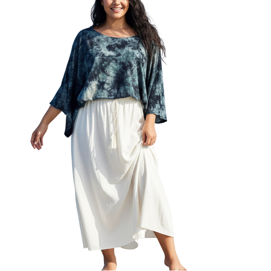 Woman standing on a wooden dock by the ocean wearing a blue tie-dye top and white skirt.