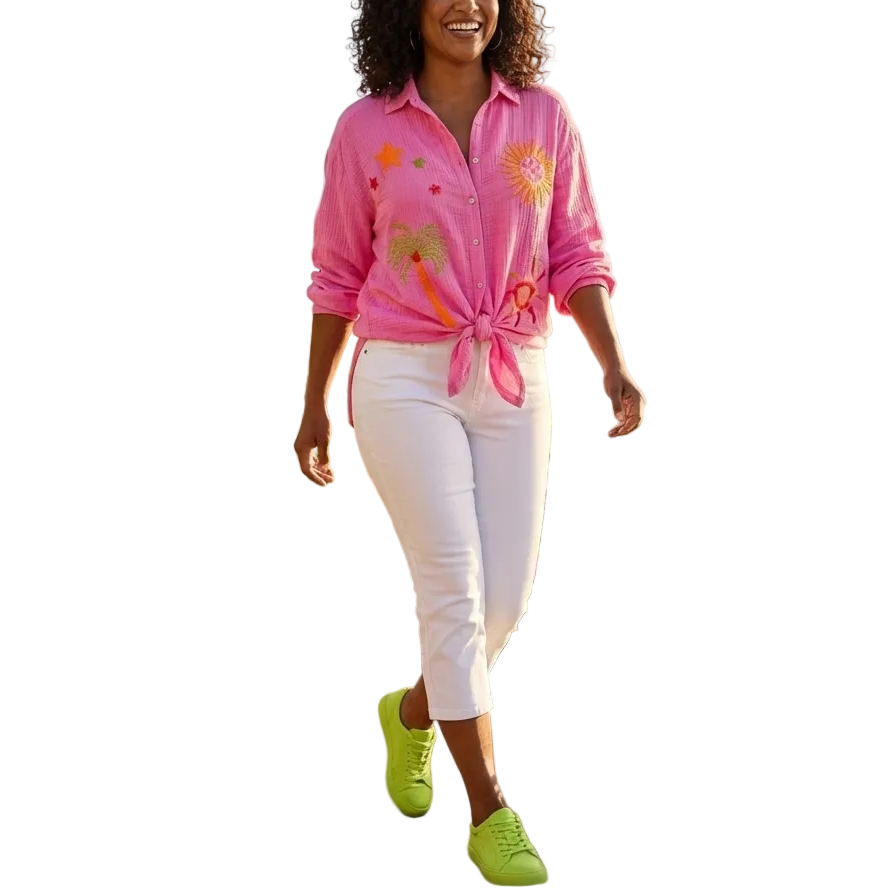 Woman in a pink shirt and white pants walking along a path with flowers and palm trees by the ocean.