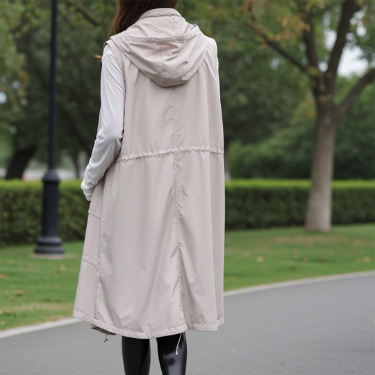 Person wearing a long beige vest over a white shirt in an outdoor setting with greenery.