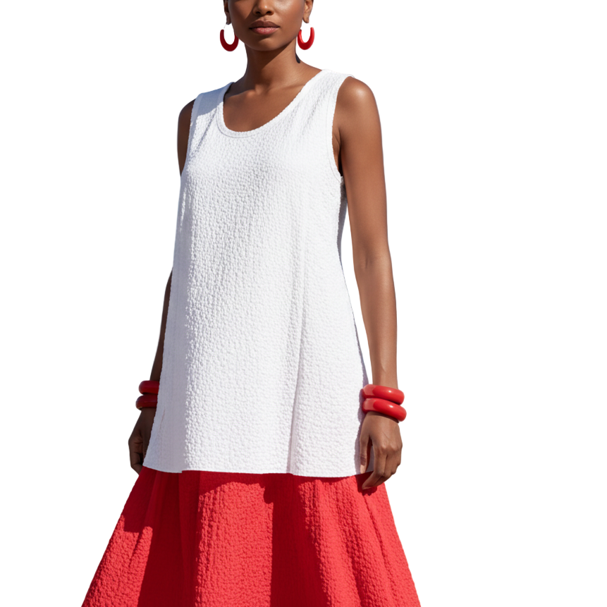 Woman in a white top and red skirt standing on a beach with blue sky and ocean.
