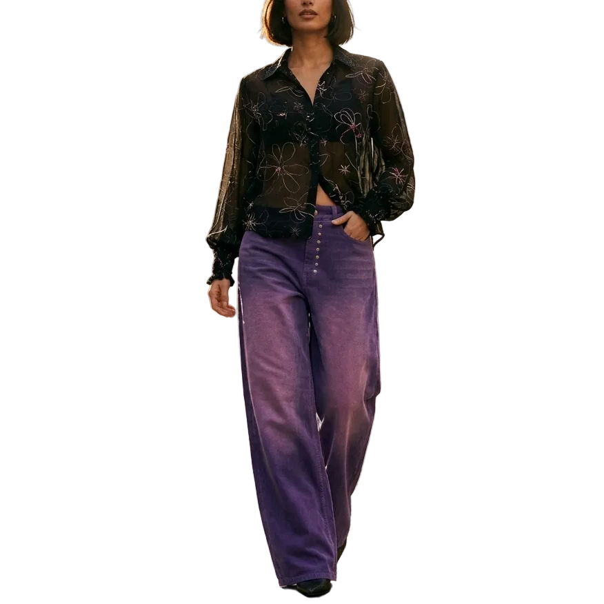 A woman stands on a cobblestone street wearing a sheer black floral long-sleeve top and high-waisted purple wide-leg trousers, with historic stone buildings and a cafe in the background.