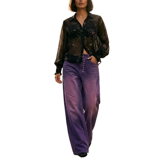 A woman stands on a cobblestone street wearing a sheer black floral long-sleeve top and high-waisted purple wide-leg trousers, with historic stone buildings and a cafe in the background.