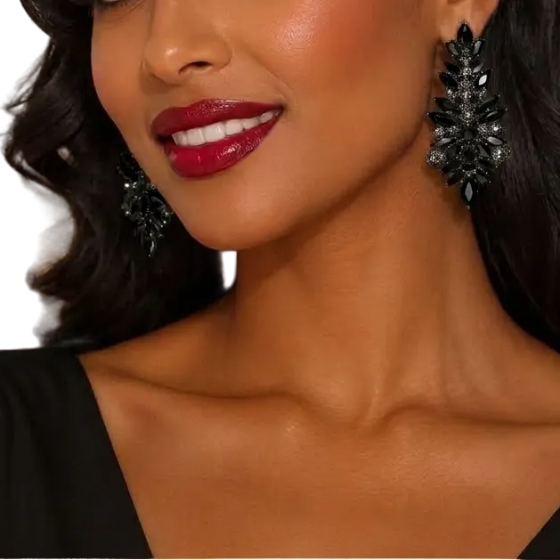 A woman with styled dark hair and red lipstick wearing large black crystal-like drop earrings and a black top, photographed in a softly lit indoor setting with blurred lights in the background.