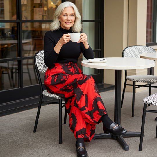 Woman sitting at a cafe table holding a cup, wearing a black top and red patterned skirt.