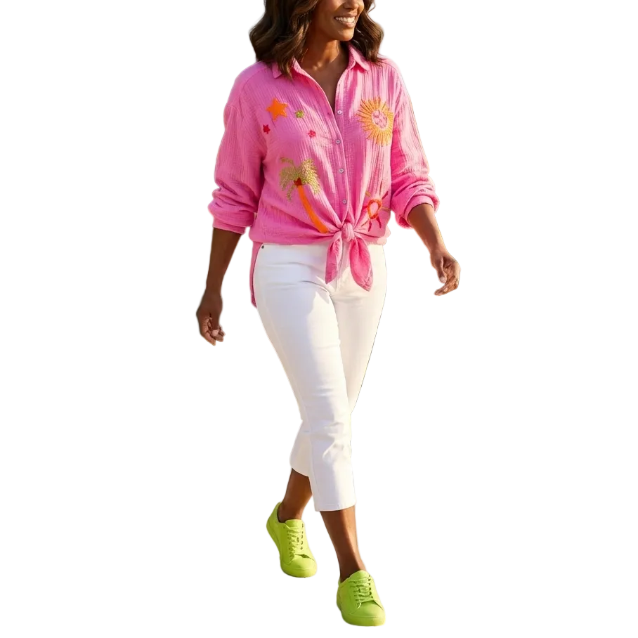 Woman in pink floral shirt and white pants walking by a pool with palm trees and flowers in the background