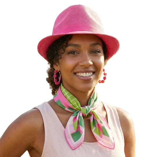 A smiling woman outdoors wearing a bright pink fedora hat, a pink and green patterned neck scarf, and a light pink sleeveless top, with blurred flowers and greenery in the background.