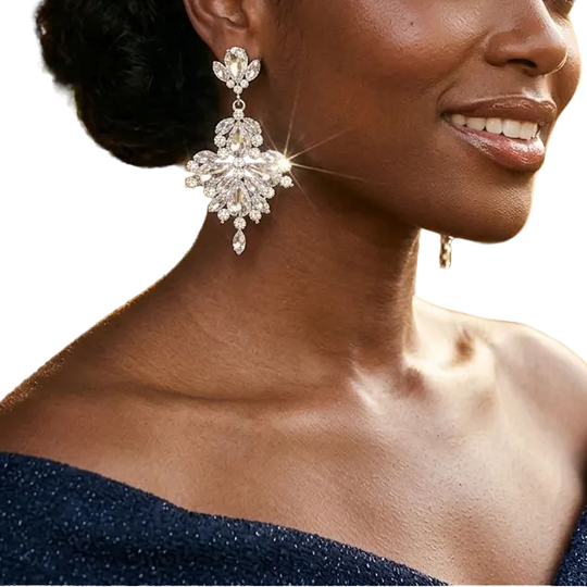 A woman with her hair in an elegant updo is shown in profile, wearing a large, sparkling silver-toned statement earring with clear gemstones. She is wearing a sparkling dark blue off-the-shoulder dress, set against a warm, softly blurred outdoor background with green foliage and golden sunlight.