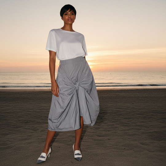 Woman wearing a white top and gray skirt standing on a beach at sunset.