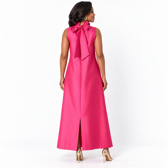 A woman modeling a sleeveless magenta pink maxi dress with a large bow at the back of the neck, shown from the rear against a white studio background.