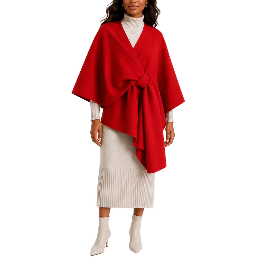 A woman modeling a bright red wrap poncho with a large front tie, worn over a cream ribbed turtleneck and a matching cream pleated midi skirt with beige ankle boots, against a plain white studio background.