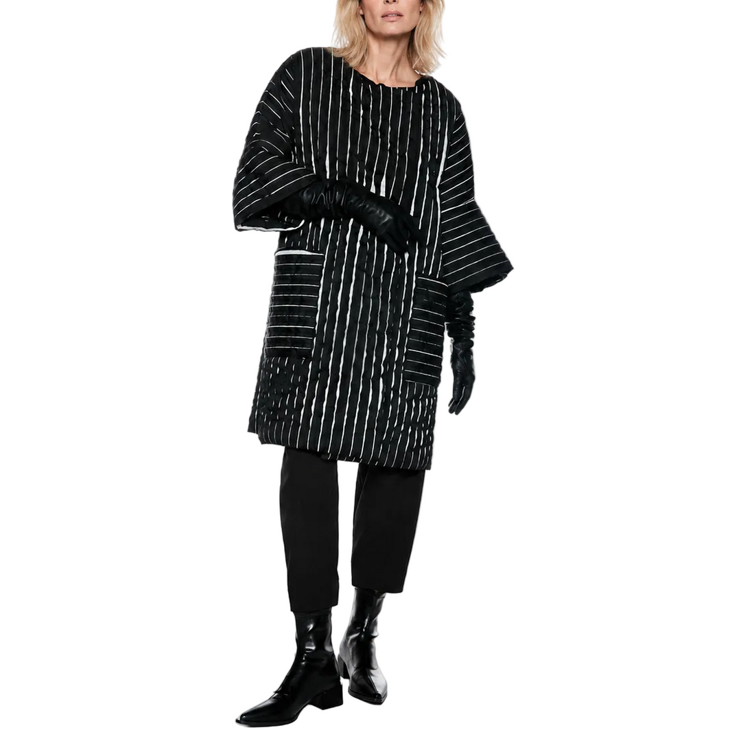 A model wearing a black and white vertically striped long-sleeve tunic top, black cropped trousers, and black ankle boots, standing against a plain white studio background.