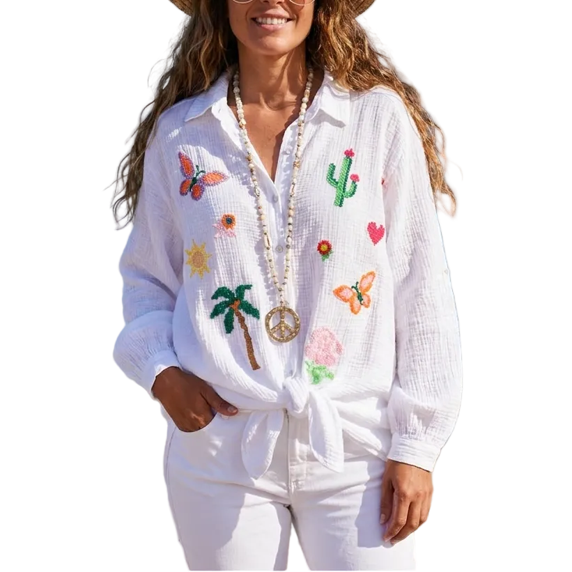 Woman on a beach wearing a white embroidered shirt and straw hat with a blue ocean and sky background.