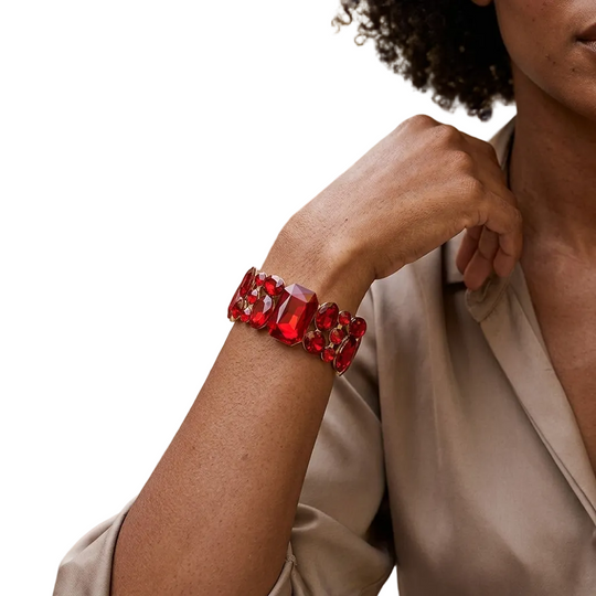 A close-up lifestyle shot of a woman wearing a chunky red beaded bracelet with gold floral accents. The background is a softly blurred outdoor patio setting with warm sunlight and greenery.