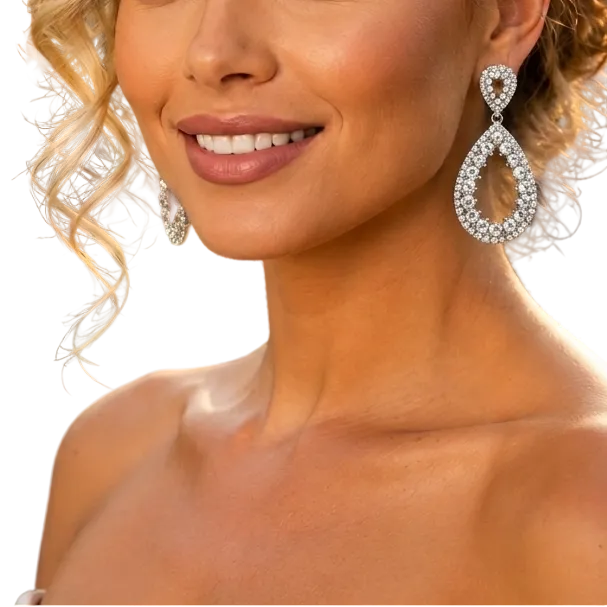 A woman with styled blonde hair and elegant makeup wearing sparkling teardrop-shaped silver and crystal drop earrings, photographed outdoors at a social event with blurred guests and string lights in the background.