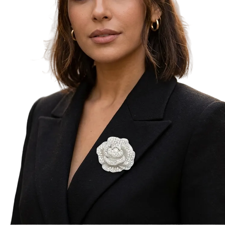 A woman with dark hair wearing a black blazer and gold hoop earrings, featuring a large silver rose brooch on her lapel, set against a softly blurred city street background.