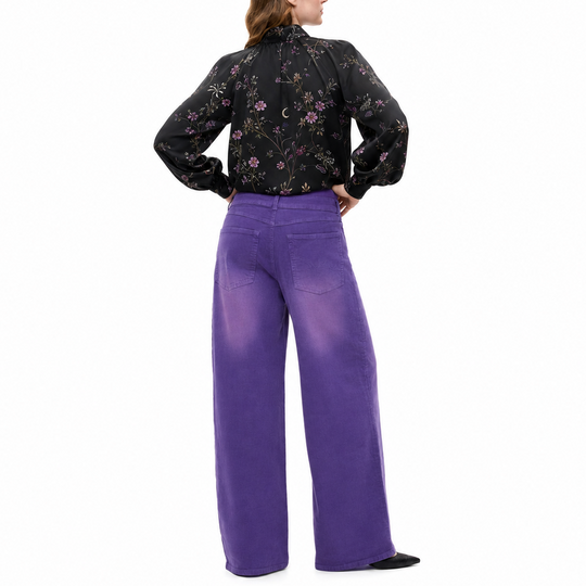 A woman shown from the back wearing a black long-sleeve blouse with a pink and green floral pattern and wide-leg purple trousers, standing against a plain white background.