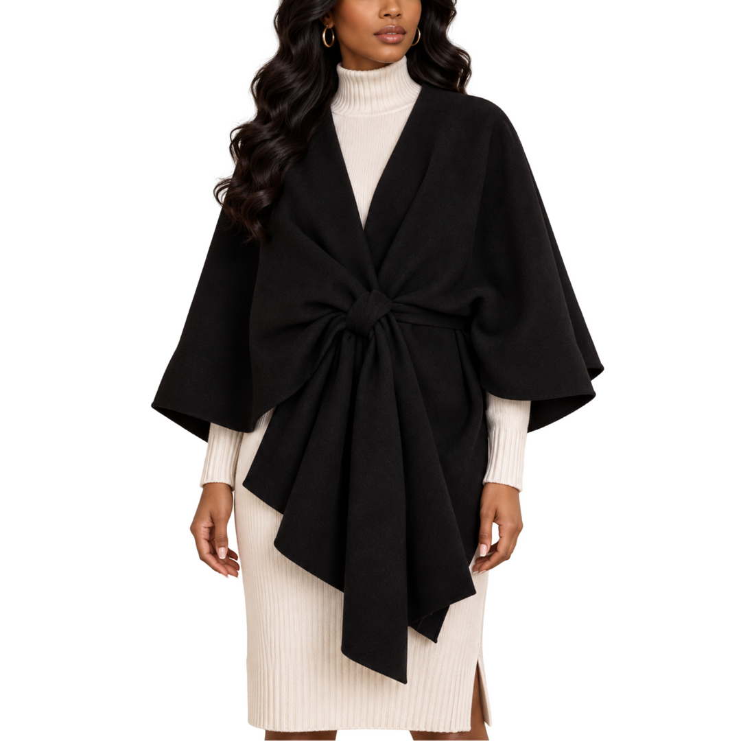 A woman modeling a black wrap poncho with a large front bow, worn over a cream ribbed turtleneck and matching cream knit skirt against a plain white background.